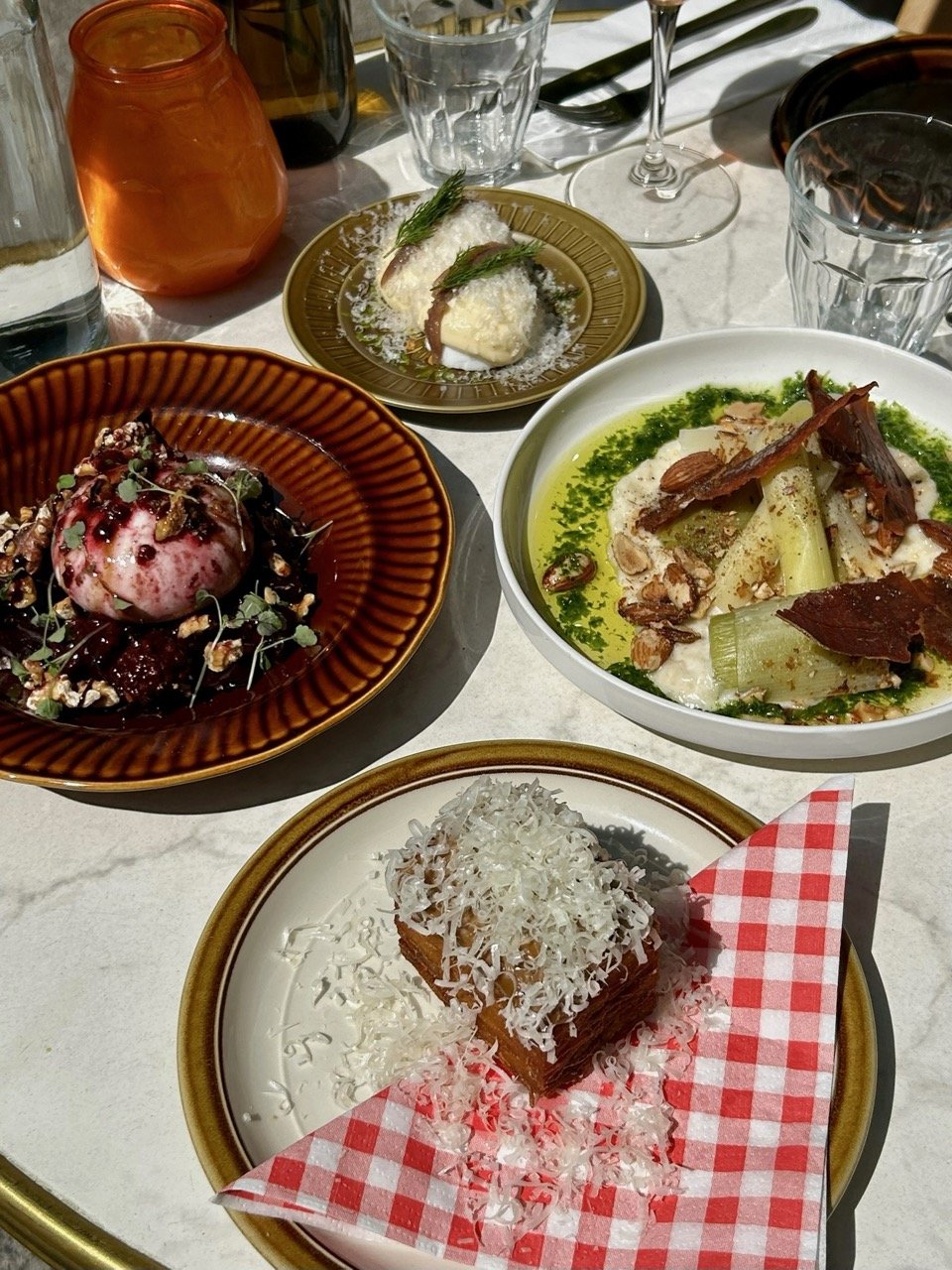 Een tafel met meerdere gerechten en drankjes, inclusief een nagerecht met witte kaas, een salade met vlees, een dessert met gesmolten kaas en Parmezaan, en kleine hapjes met groenten en dressings.
