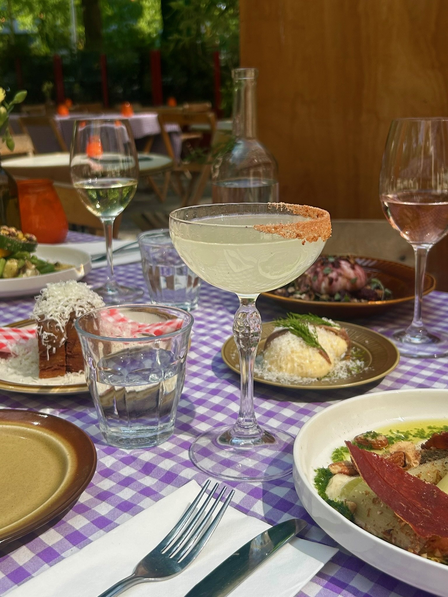 Gedeelde tafel met diverse gerechten en drankjes op een tafelkleed met een paar glazen wijn, een cocktail, water en een fles water. Er is een bord met salade en een dessert zichtbaar.