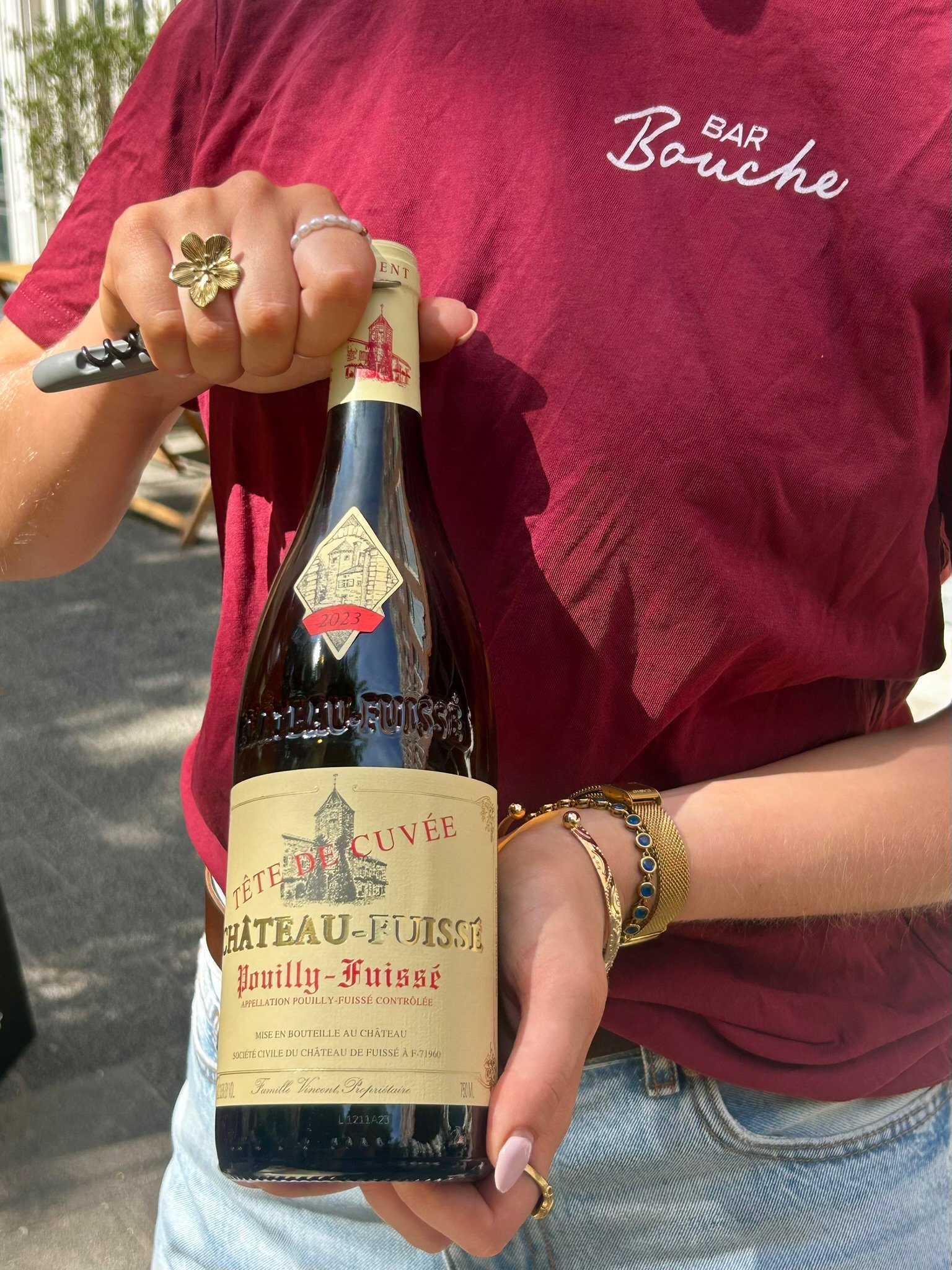 Persoon houdt een fles rode wijn vast met een rode T-shirt met 'Bar Bouche' erop.